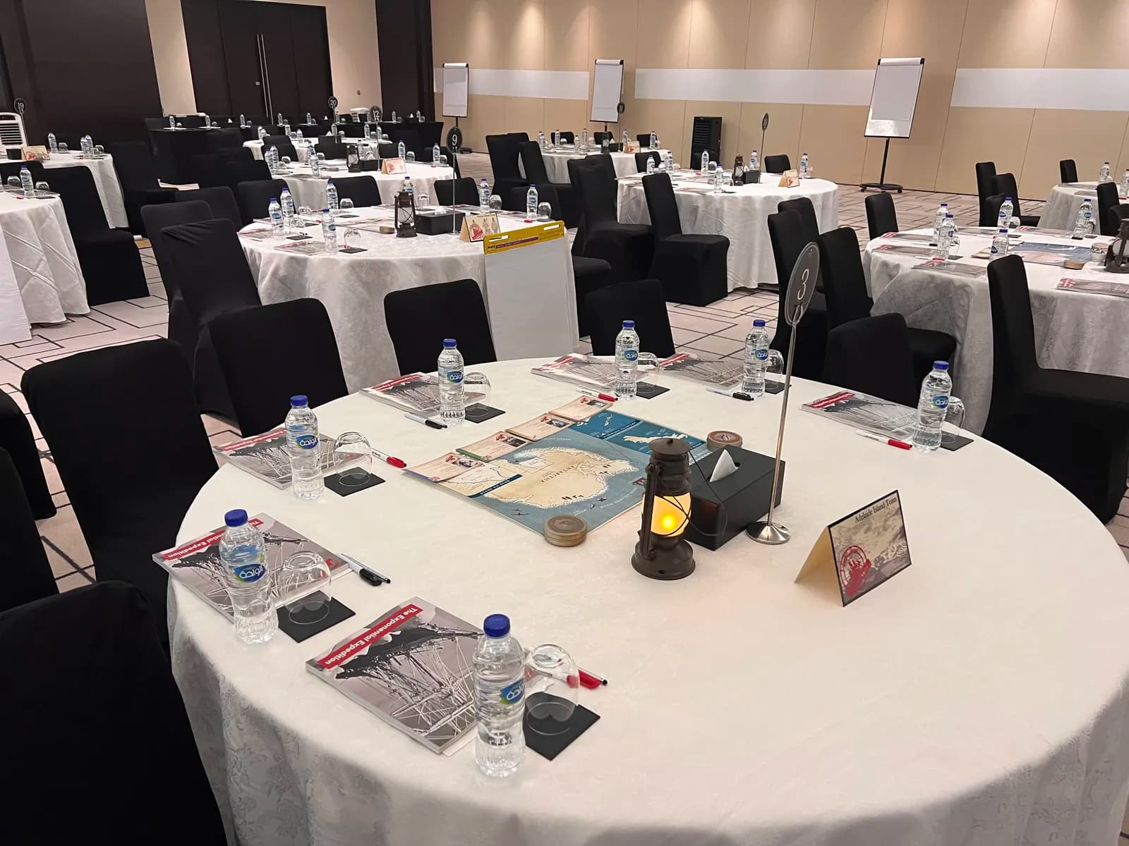 Lead the Endurance experience room setup — multiple round tables with Shackleton expedition workbooks, Antarctica maps, lanterns, and leadership materials ready for participants
